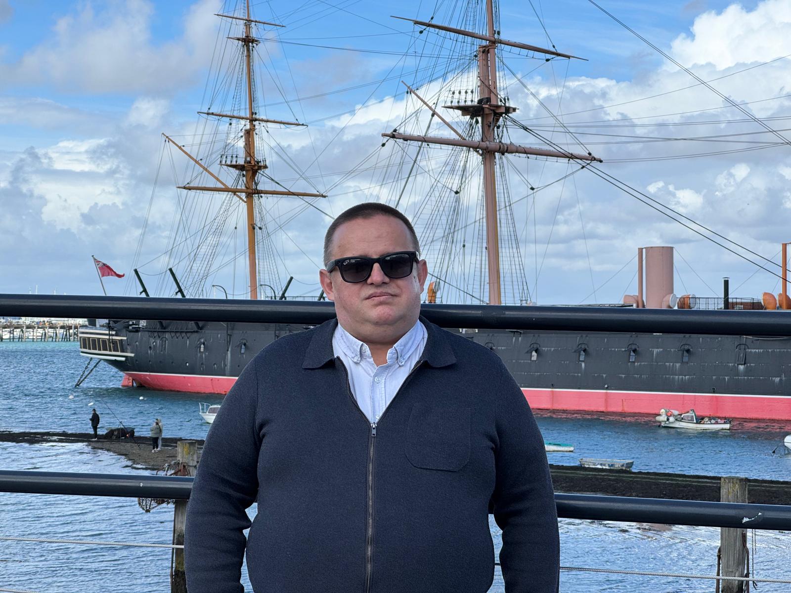 Adrian Tamasan standing by the water in Portsmouth, with the historic ship HMS Warrior behind him